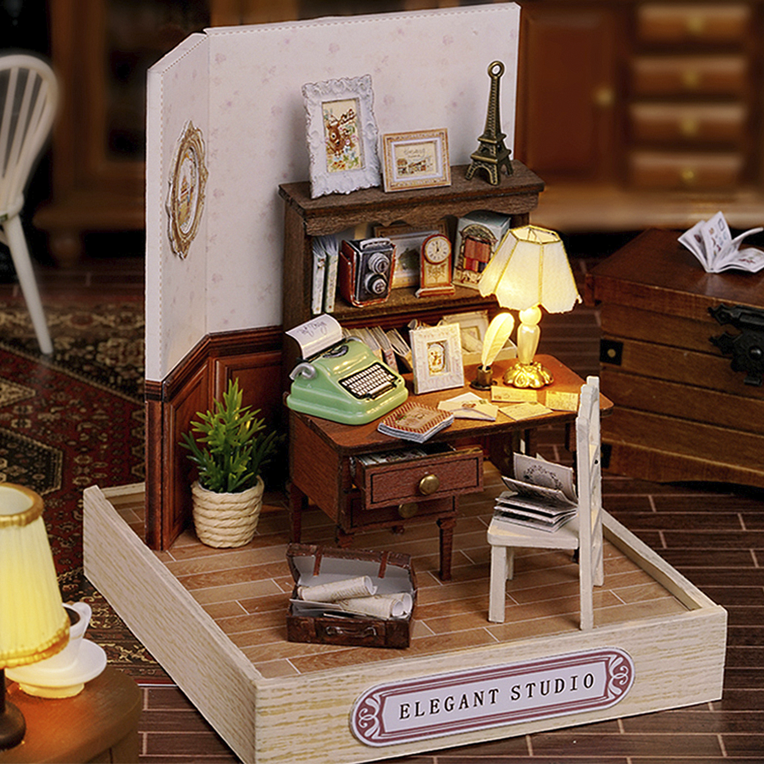 Studio elegante in miniatura con scrivania, macchina da scrivere, lampada e piccola replica della Torre Eiffel. Include una pianta in vaso, sedia e valigia aperta.