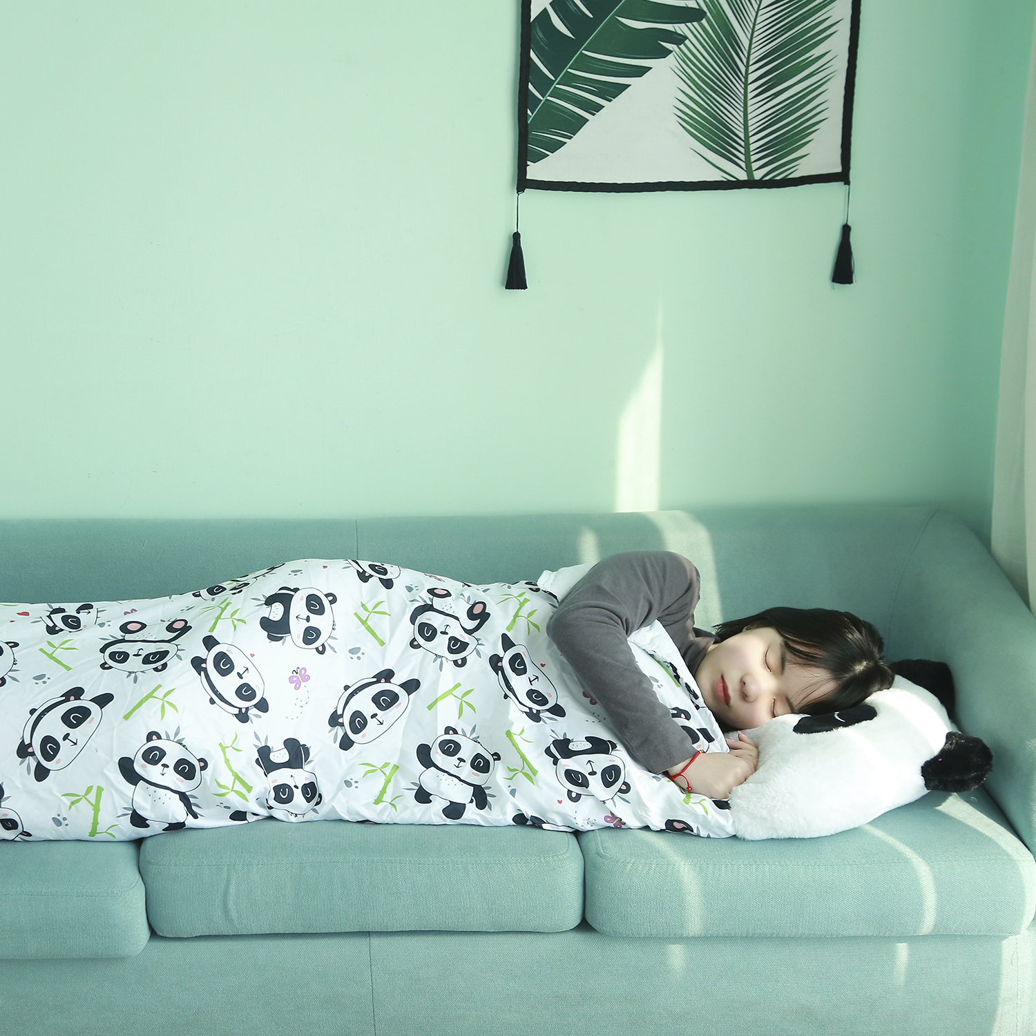 Una persona dorme su un divano sotto una coperta a tema panda. Viene utilizzato un cuscino panda. Lo sfondo è una parete con un'immagine di piante.