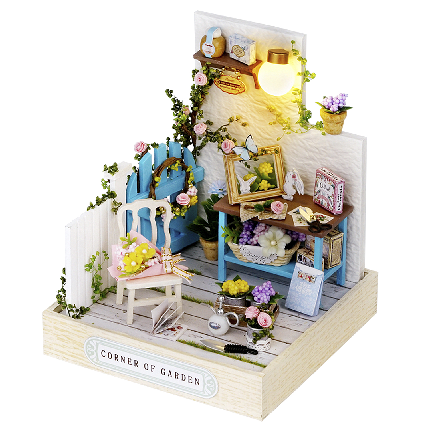 Scena di giardino in miniatura con una recinzione blu, una sedia, un tavolo, fiori e una lampada. La scena è etichettata 'Corner of Garden'.
