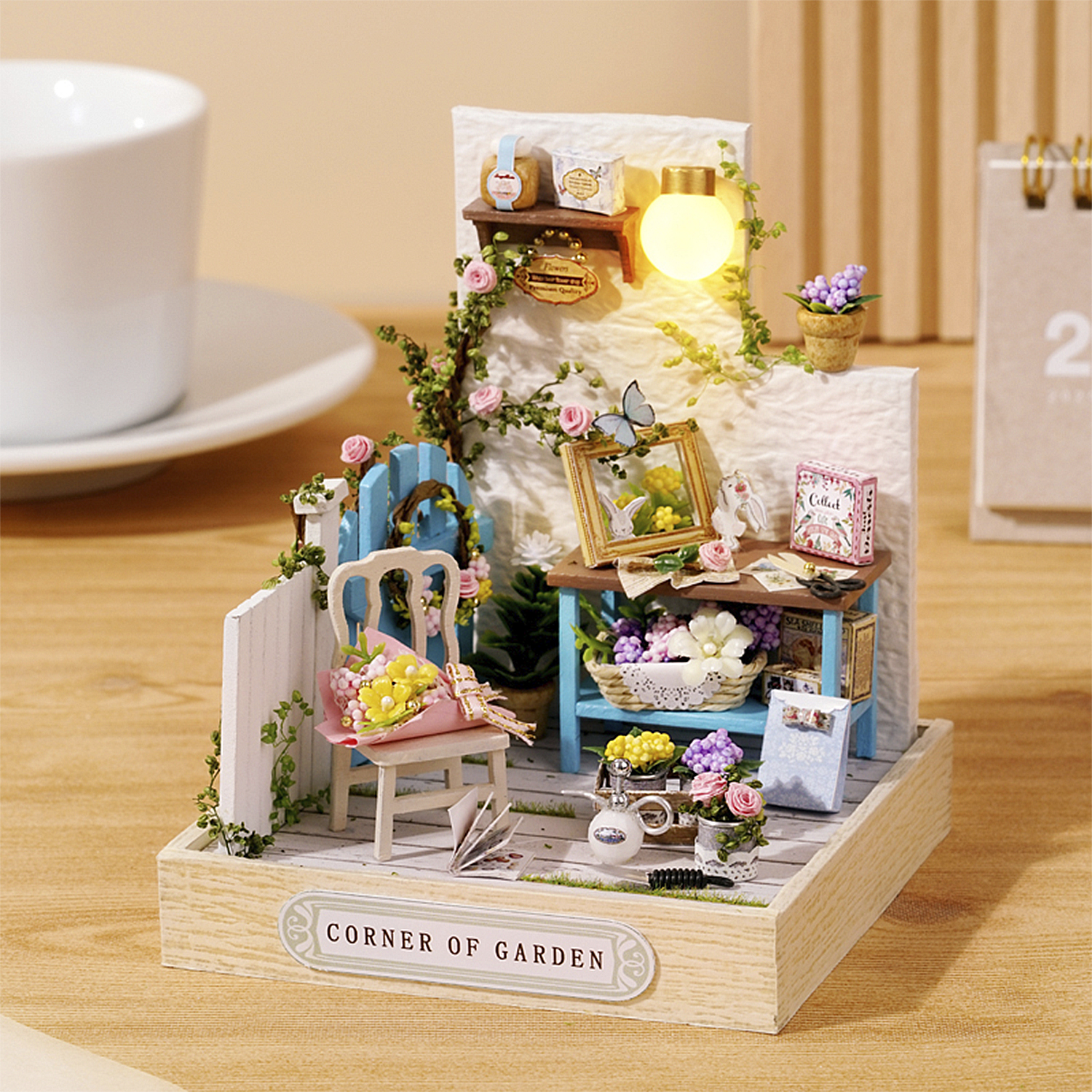 Scena di giardino in miniatura con fiori, un tavolo e una sedia. Un bouquet è sulla sedia. C'è una luce e un calendario.