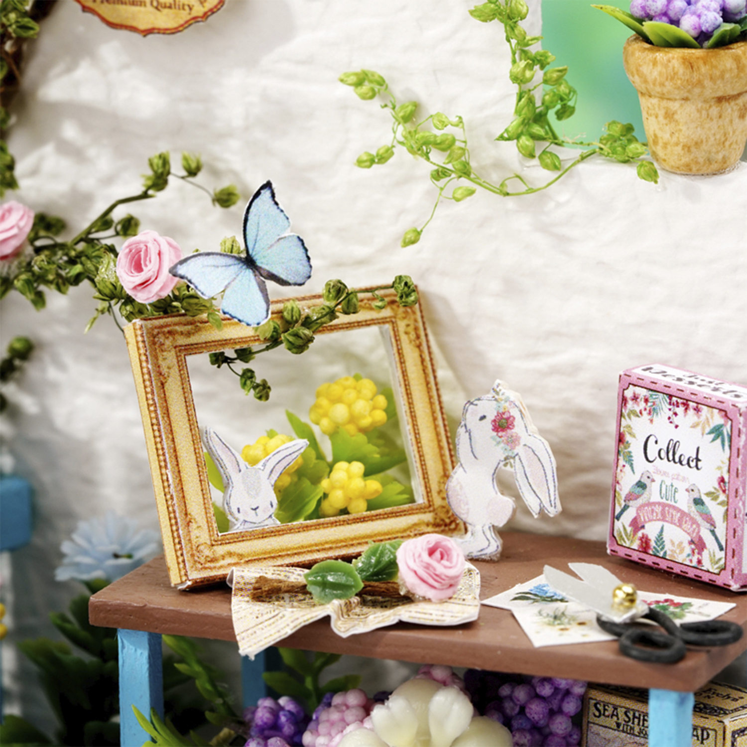 Scena in miniatura con cornice, farfalla, figure di coniglietti, fiori, tavolino e scatola. I colori tenui dominano.