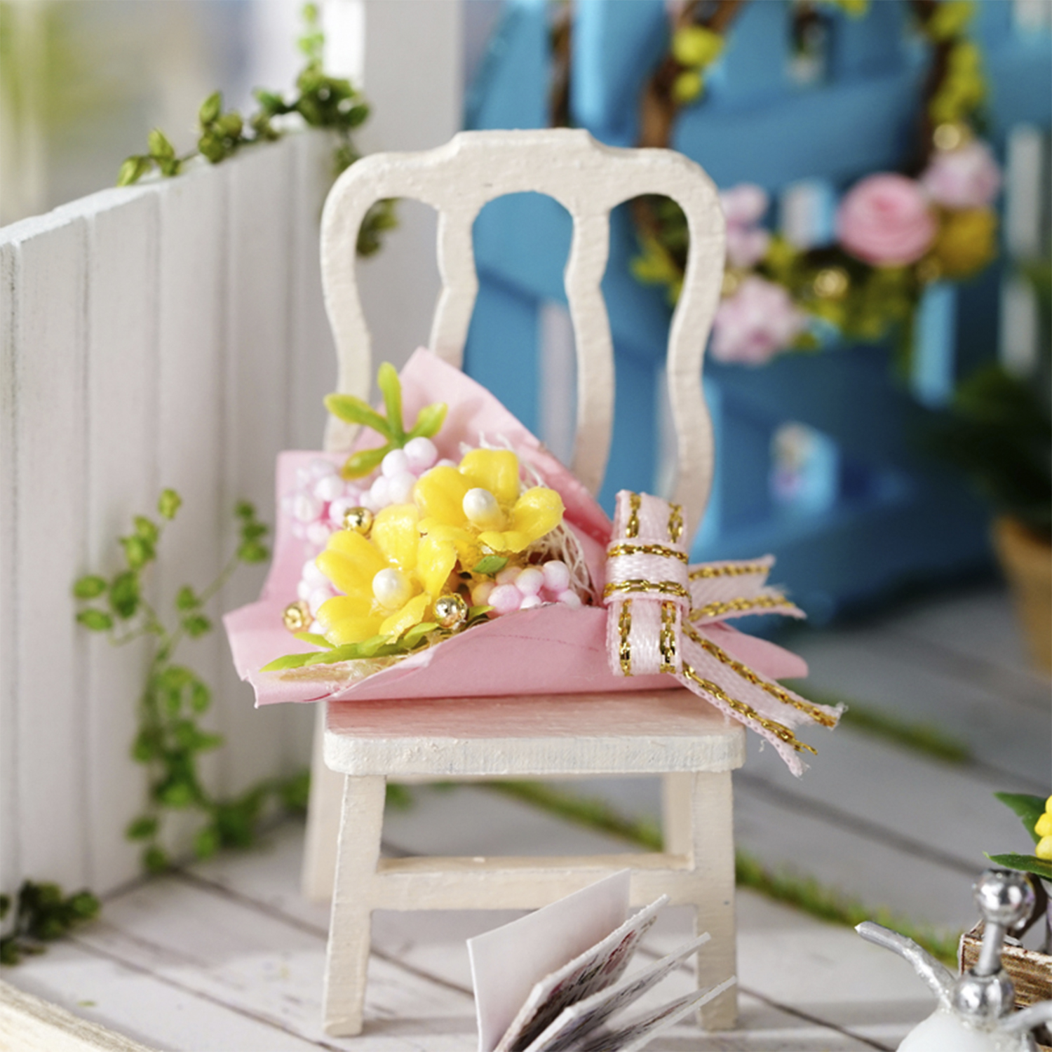 Una scena in miniatura con una sedia bianca che tiene un mazzo di fiori gialli. Incarto rosa e fiocco dorato. Sfondo blu.