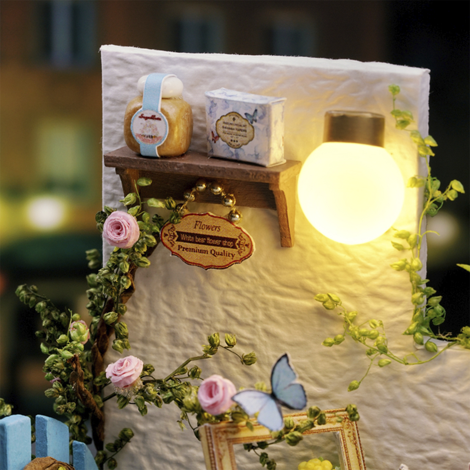 Scena in miniatura con una mensola che contiene oggetti, una lampadina accesa, rose, edera e una farfalla blu. La scena assomiglia a una casa delle bambole.