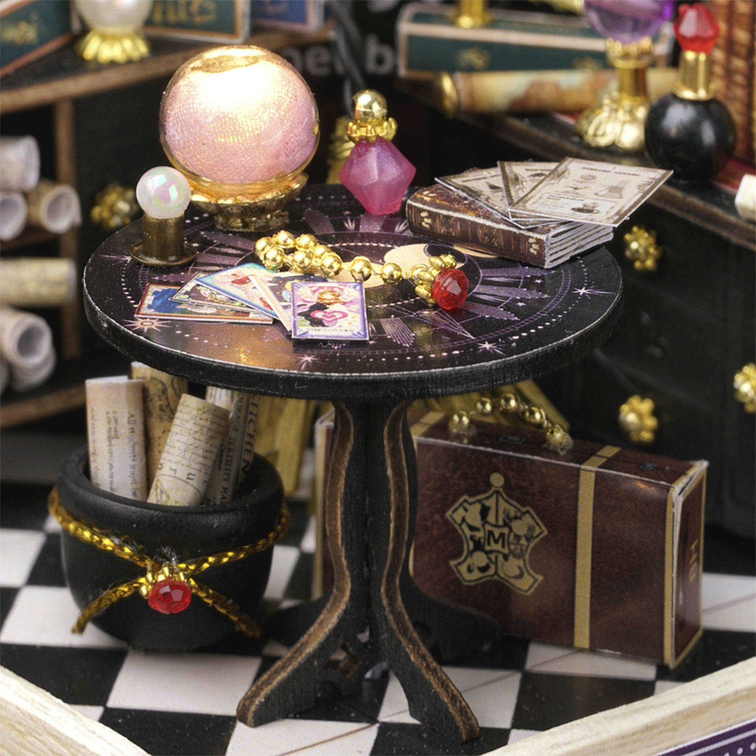 Una scena in miniatura con una sfera di cristallo, carte, libri e un calderone su un tavolo. Presente un piccolo baule.