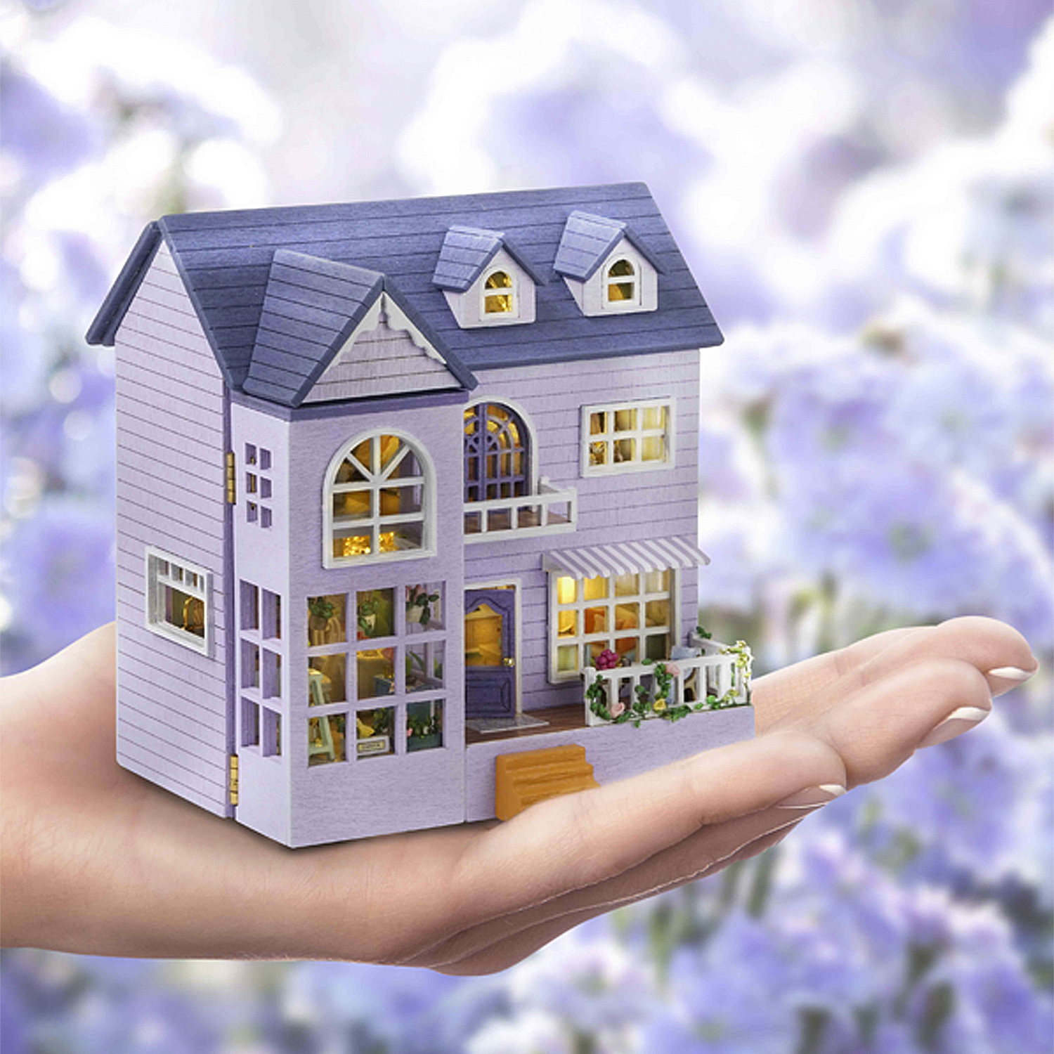 Una casa lavanda in miniatura con tetto blu è tenuta in mano. La casa ha finestre illuminate e un piccolo portico.