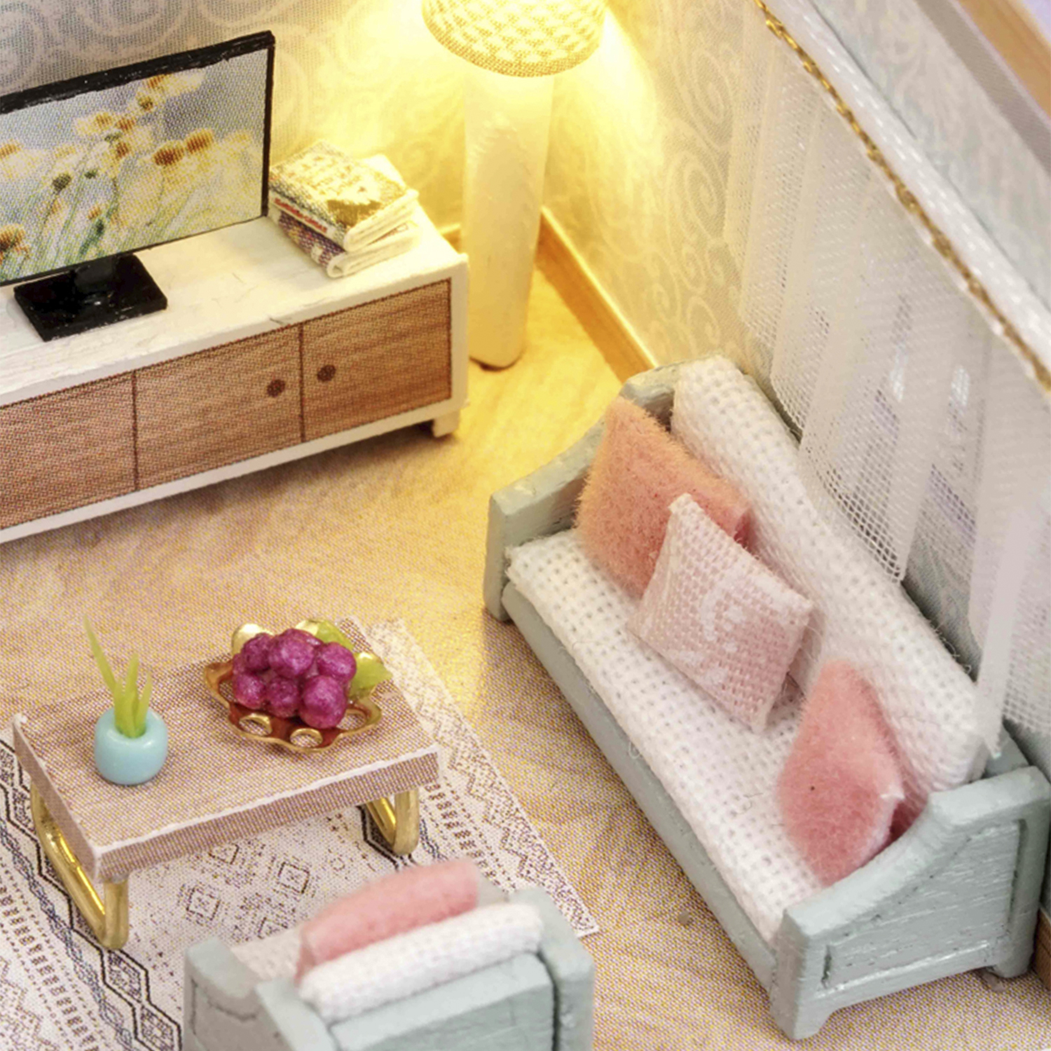 Soggiorno in miniatura con divano, tavolino con frutta, TV e lampada. Cuscini rosa e bianchi. Mobili in legno.