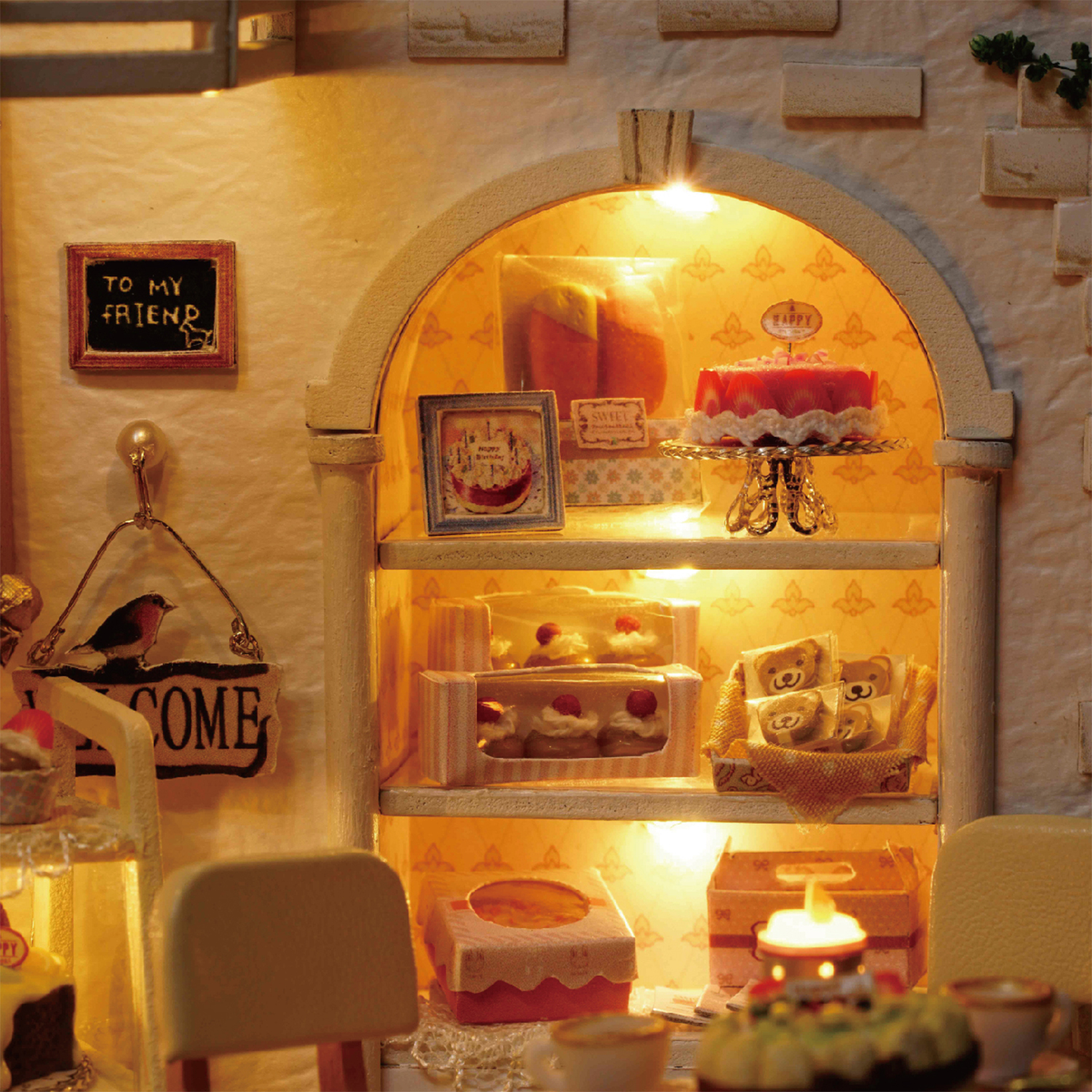 Miniaturbäckerei mit Kuchen, Gebäck und einem Schild 'An meinen Freund'. Beleuchtet von warmen Lichtern, was eine gemütliche Atmosphäre schafft.