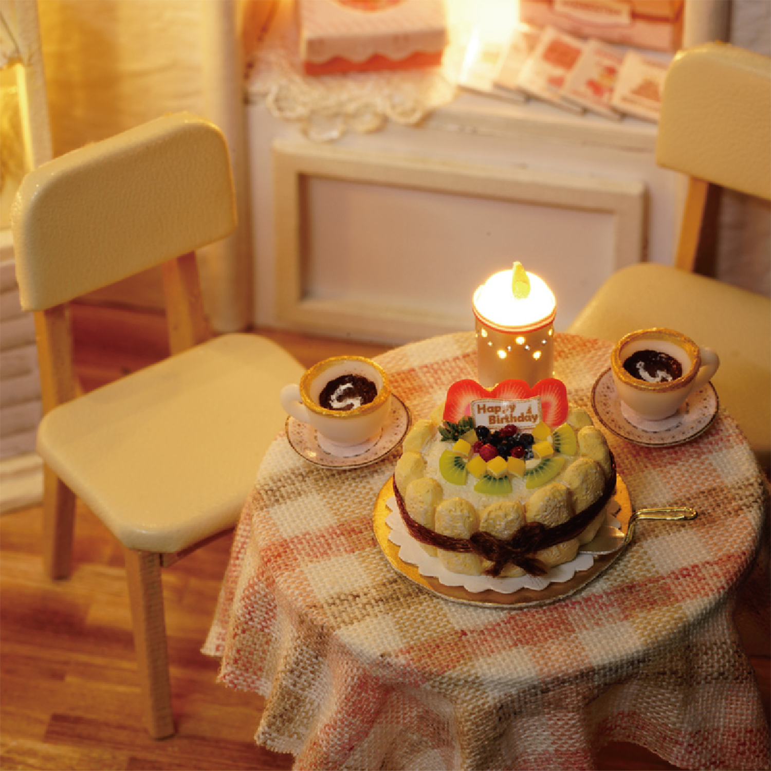 Miniatur-Geburtstagsszene. Kuchen mit Obst, zwei Kaffeetassen, brennende Kerze und zwei Stühle um einen Tisch mit gemustertem Tuch.