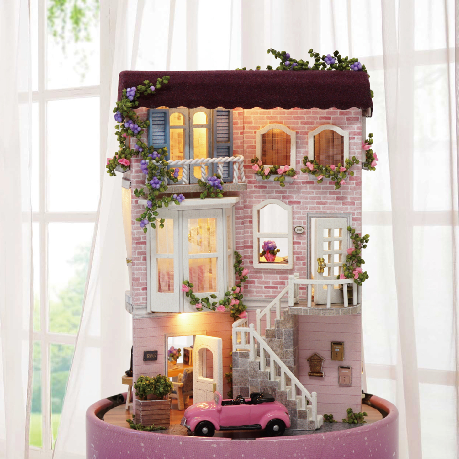 Una casa rosa in miniatura con auto, fiori e facciata in mattoni. La casa ha più piani e luci all'interno.