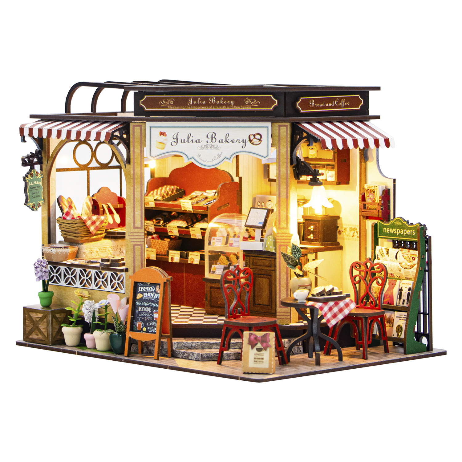 Una panetteria in miniatura con tenda a strisce rosse e bianche, esposizione di pane, tavolini, sedie e un'edicola.