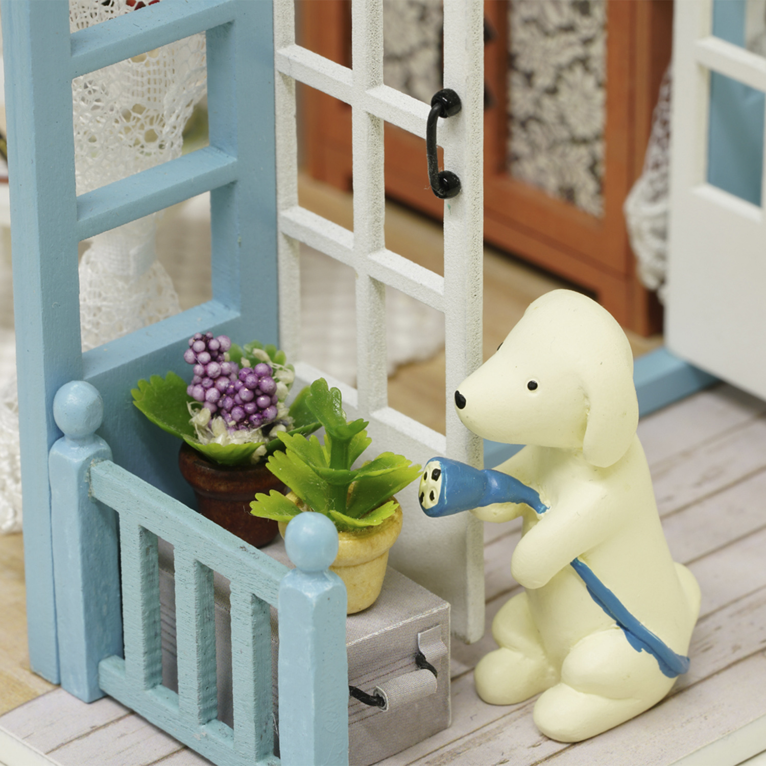 Ein Spielzeughund gießt Pflanzen auf einem Miniatur-Balkon. Der Hund ist weiß, der Balkon blau. Töpfe mit Pflanzen und eine Holztür.