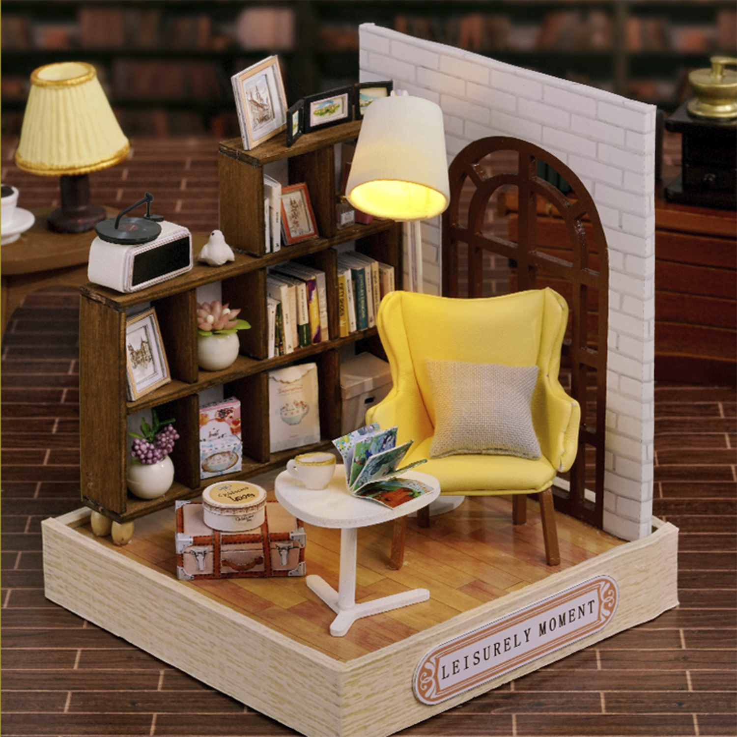 Stanza in miniatura con sedia gialla, libri, lampada e tavolino. Un cartello dice 'LEISURELY MOMENT'.