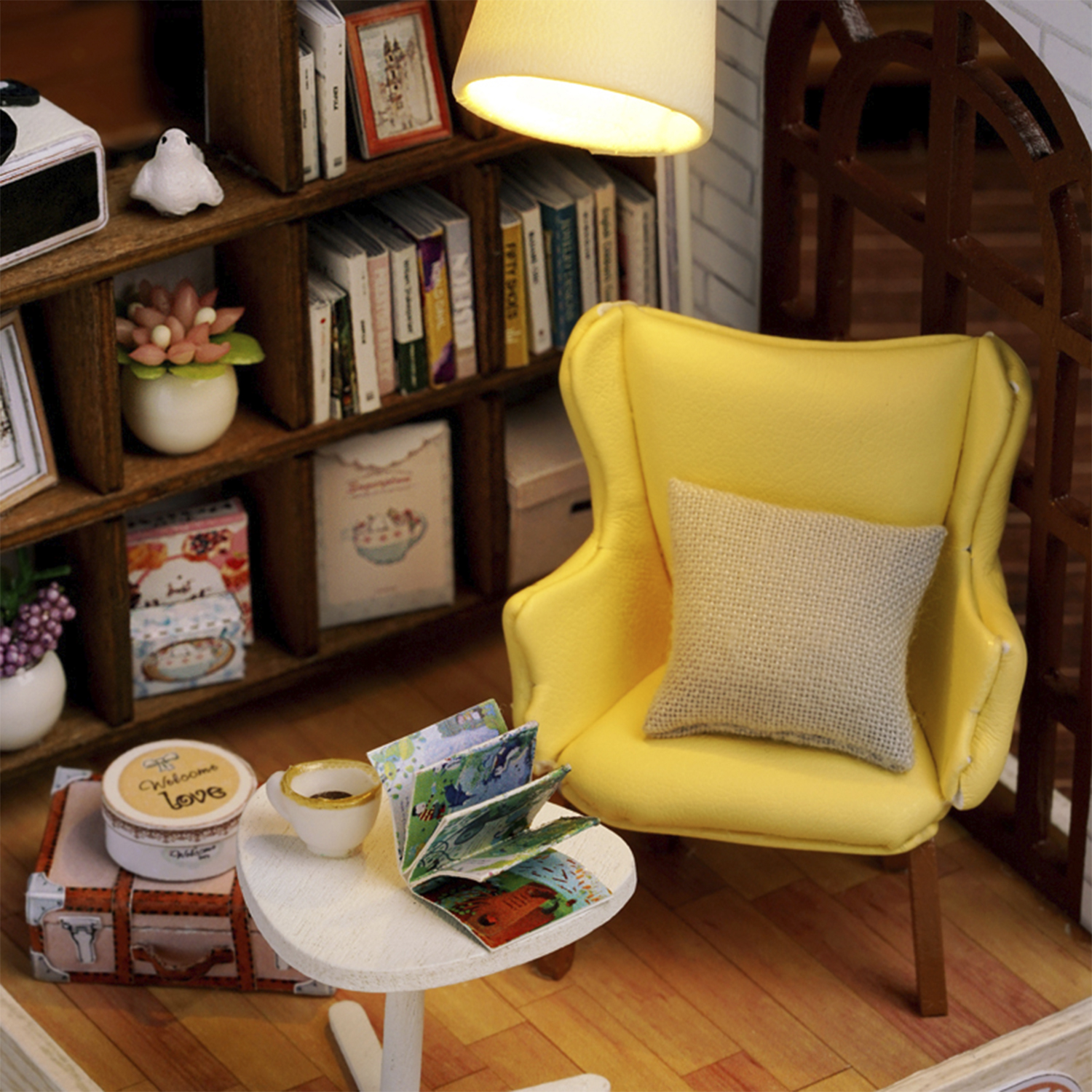 Stanza in miniatura con sedia gialla, libri, tavolino con tazza di caffè e libro aperto. Libreria e lampada presenti.