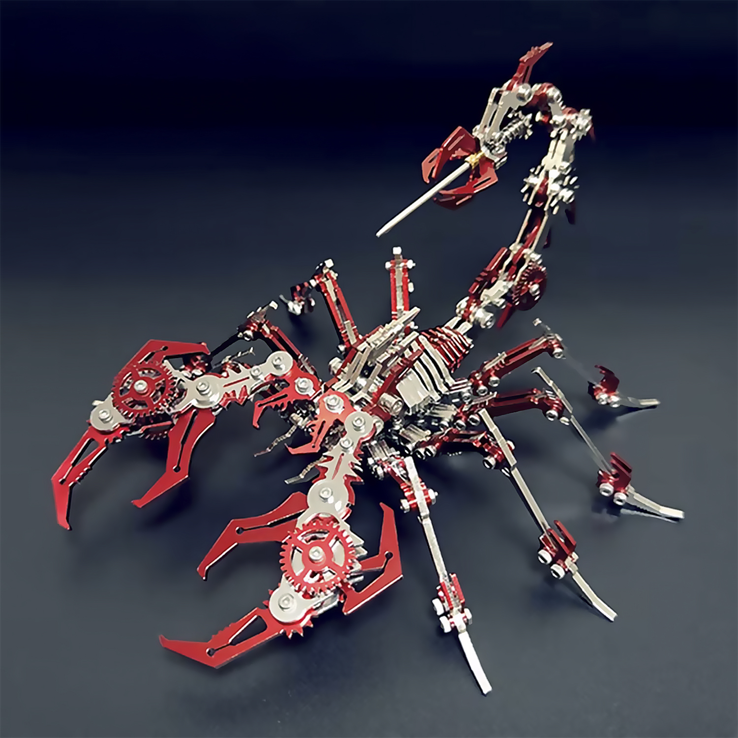 Un modello di scorpione metallico con parti rosse e argento. Ha ingranaggi, chele e una coda curva. Sfondo scuro e riflettente.