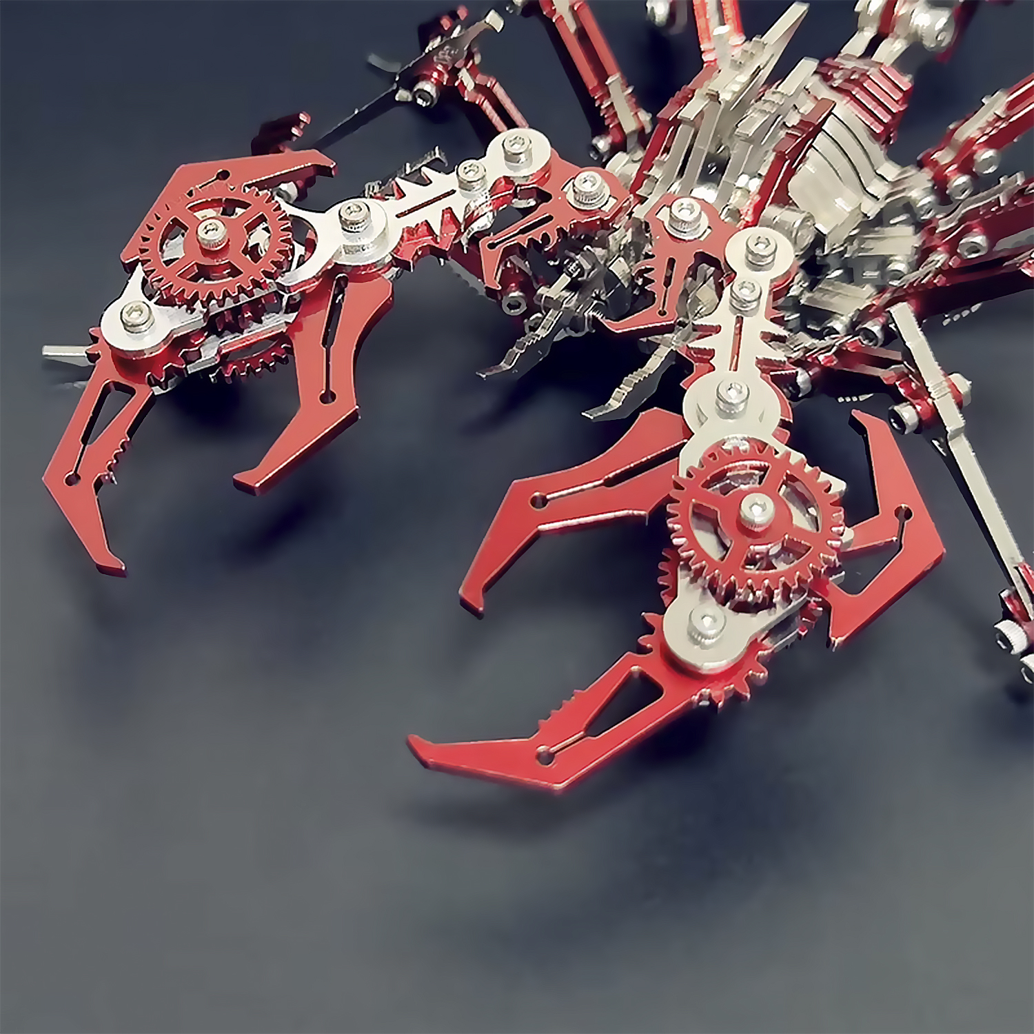 Primo piano di un modello di scorpione metallico. Presenta parti rosse e argentate, ingranaggi e intricati dettagli meccanici su sfondo scuro.