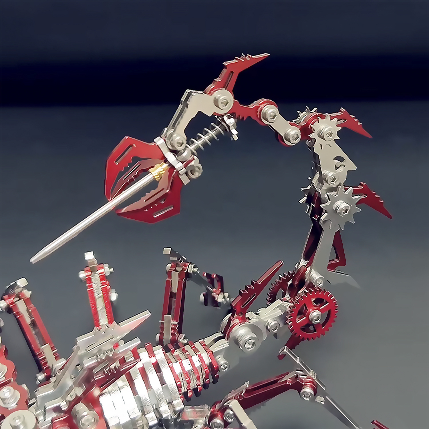 Un modello di scorpione in metallo con parti rosse e argento. Ha ingranaggi e artigli meccanici. Lo sfondo è grigio.