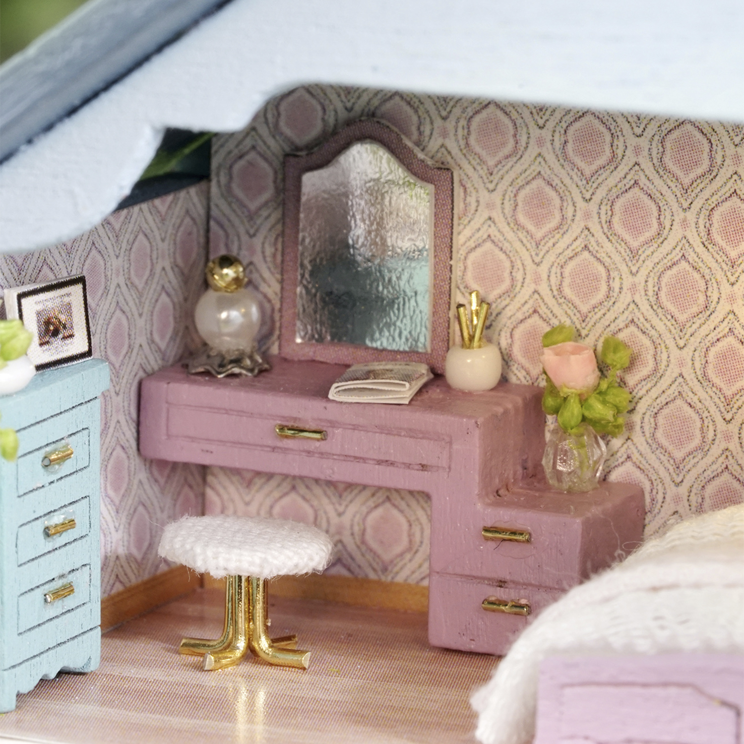 Camera da letto in miniatura con toeletta rosa, specchio, sgabello bianco e armadio blu. Carta da parati con motivo rosa. Un letto è a destra.