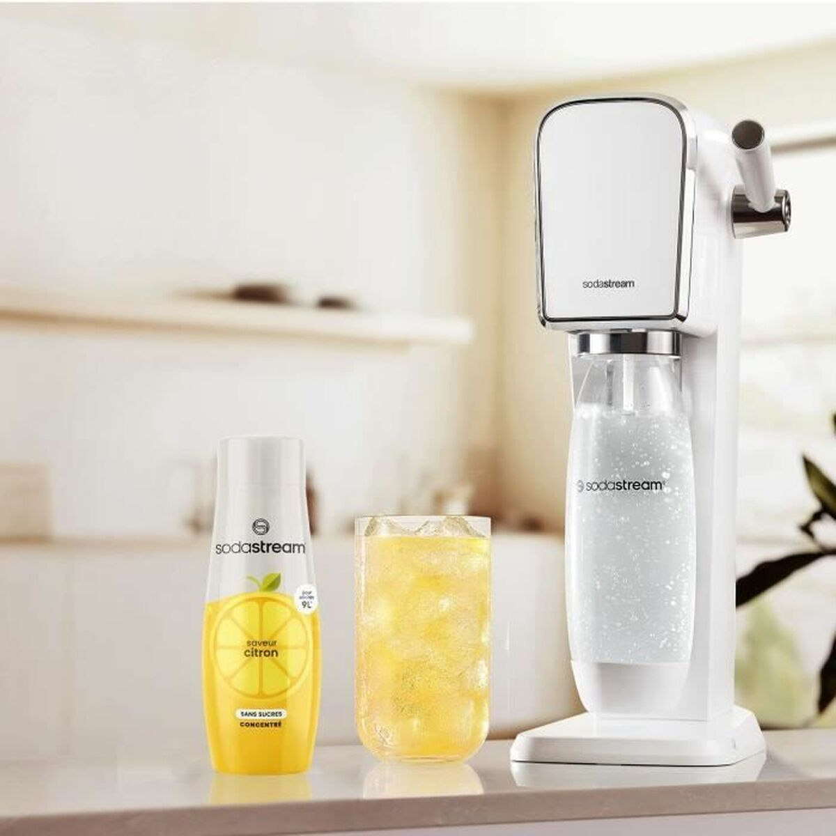 Eine weiße SodaStream-Maschine mit einer Flasche Sprudelwasser neben einem gelben Sirup mit Zitronengeschmack und einem Glas gelbem Getränk.