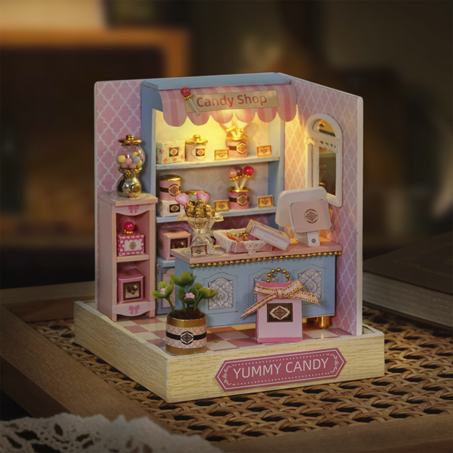 Miniatur-Süßwarenladen mit rosa Wänden und blauem Tresen. Regale halten Leckereien, einen Kaugummiautomaten und eine kleine Pflanze. Yummy Candy Schild.
