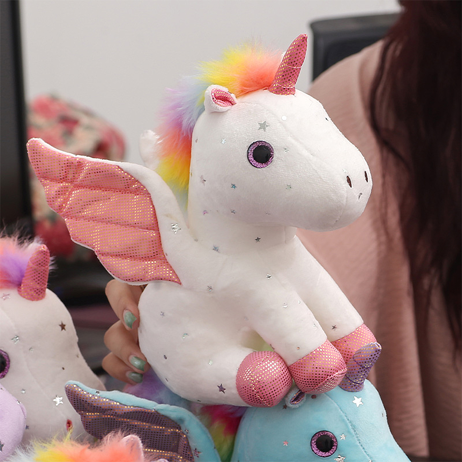 Un unicorno di peluche bianco con criniera arcobaleno, ali e zoccoli rosa, tenuto da una persona. Altri unicorni sullo sfondo.