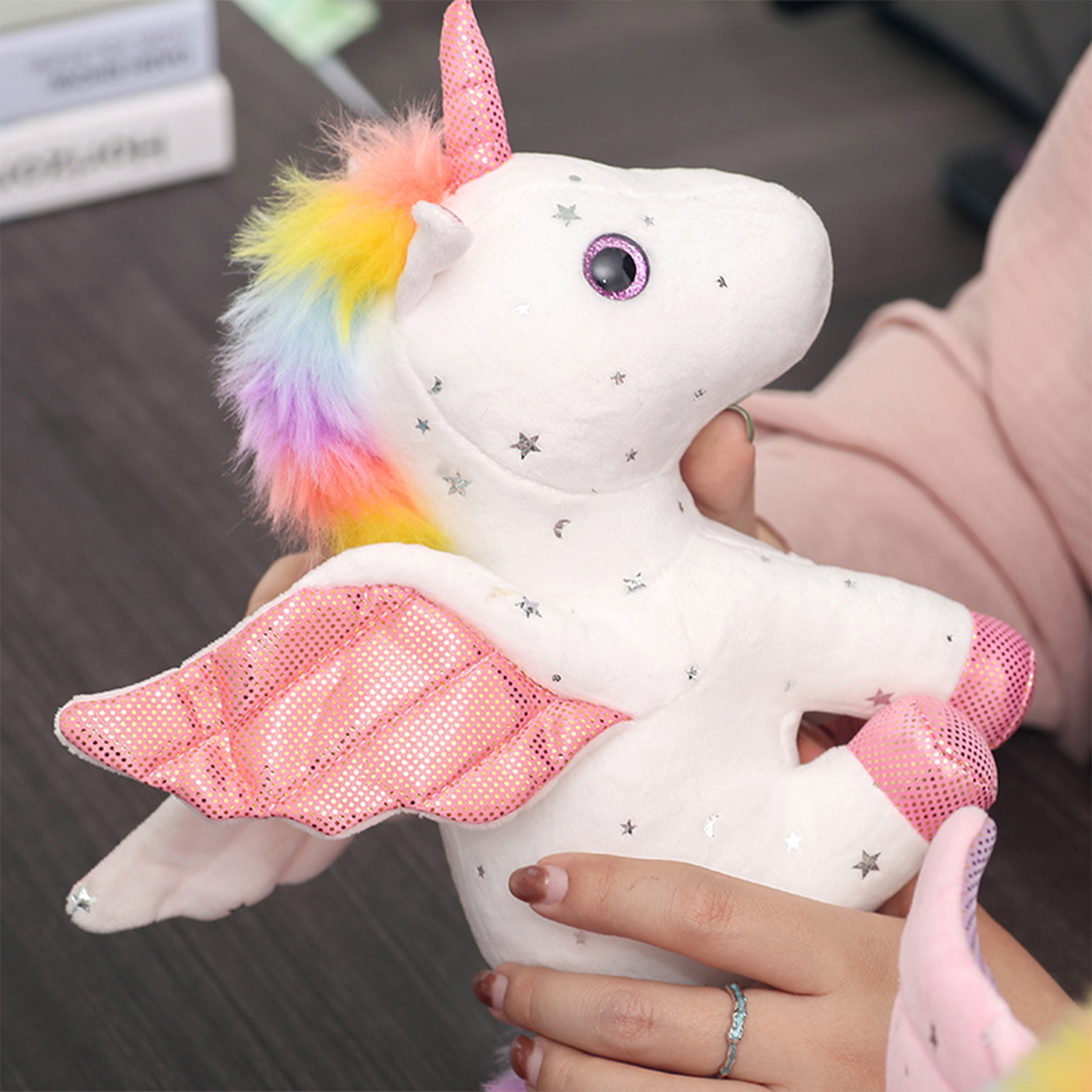 Un peluche a forma di unicorno bianco con ali e corno rosa, tenuto in mano da una persona. L'unicorno ha una criniera arcobaleno e dettagli a stella.