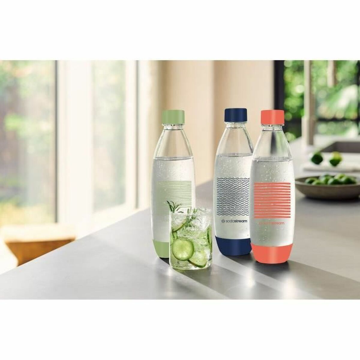 Tre bottiglie di soda con tappi e motivi colorati, un bicchiere di bevanda al cetriolo e una ciotola di lime su un bancone.