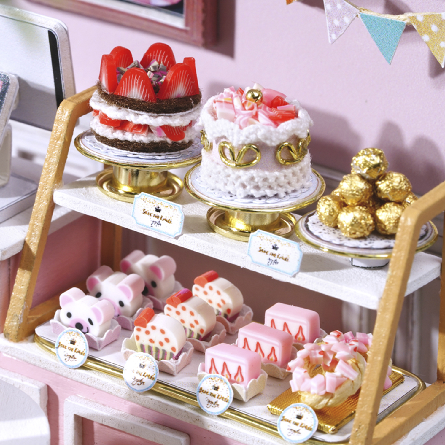 Miniature torte e dolci esposti. Include torta alle fragole, torte rosa, ciambelle e palline di cioccolato dorate. Sfondo rosa del negozio.