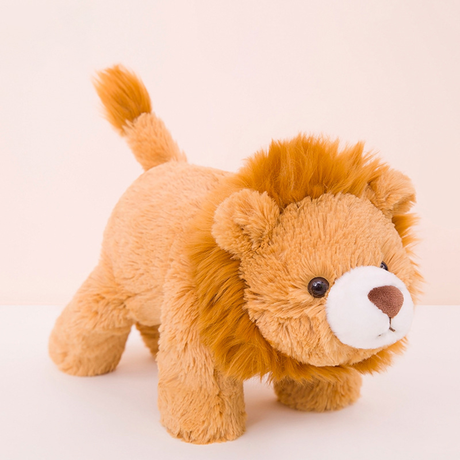 Un leone di peluche con criniera e corpo arancione, occhi e naso marroni, e una coda, è in piedi su una superficie bianca contro uno sfondo rosa.
