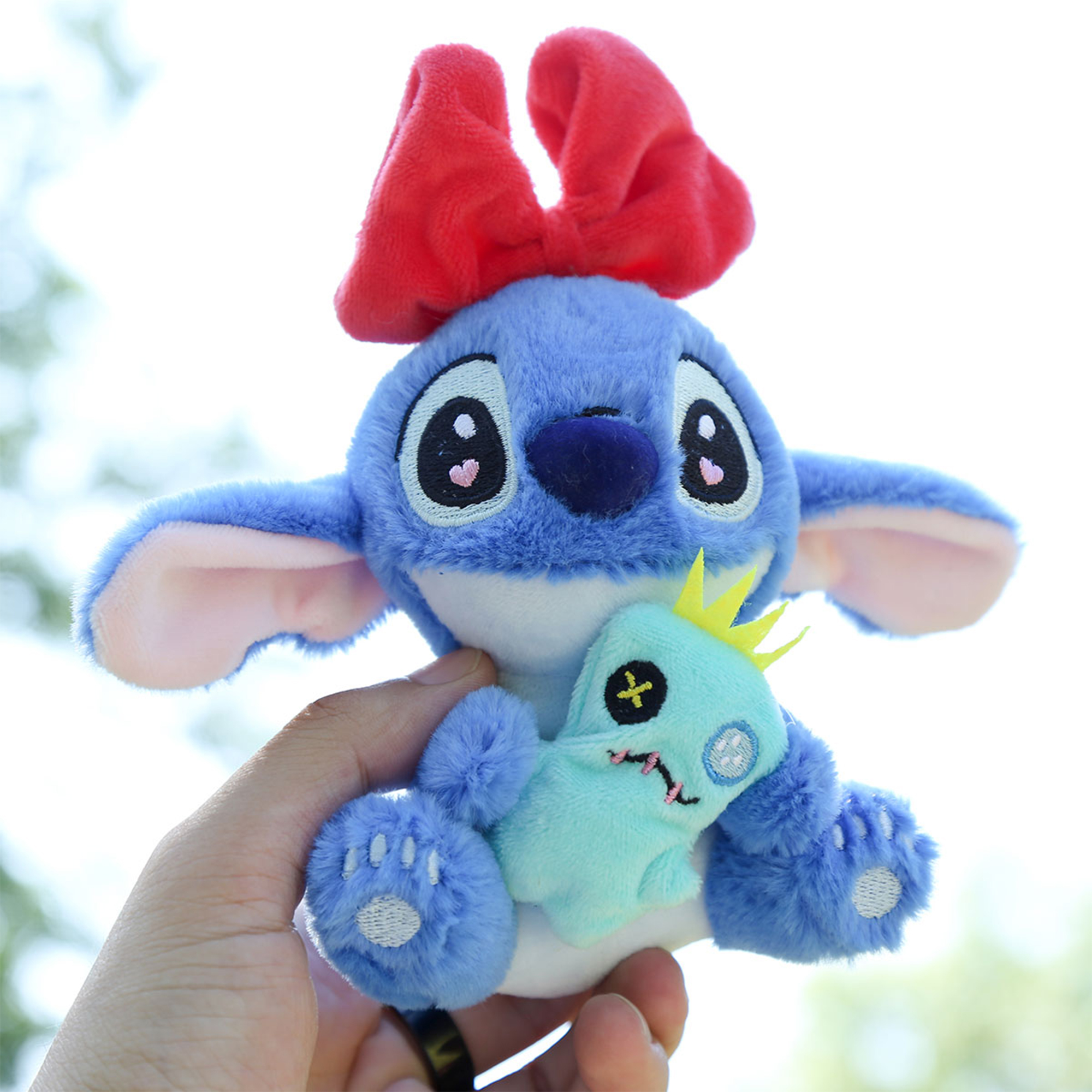 Una mano tiene un peluche blu di Stitch con un fiocco rosso e un altro piccolo peluche. Lo sfondo è luminoso.