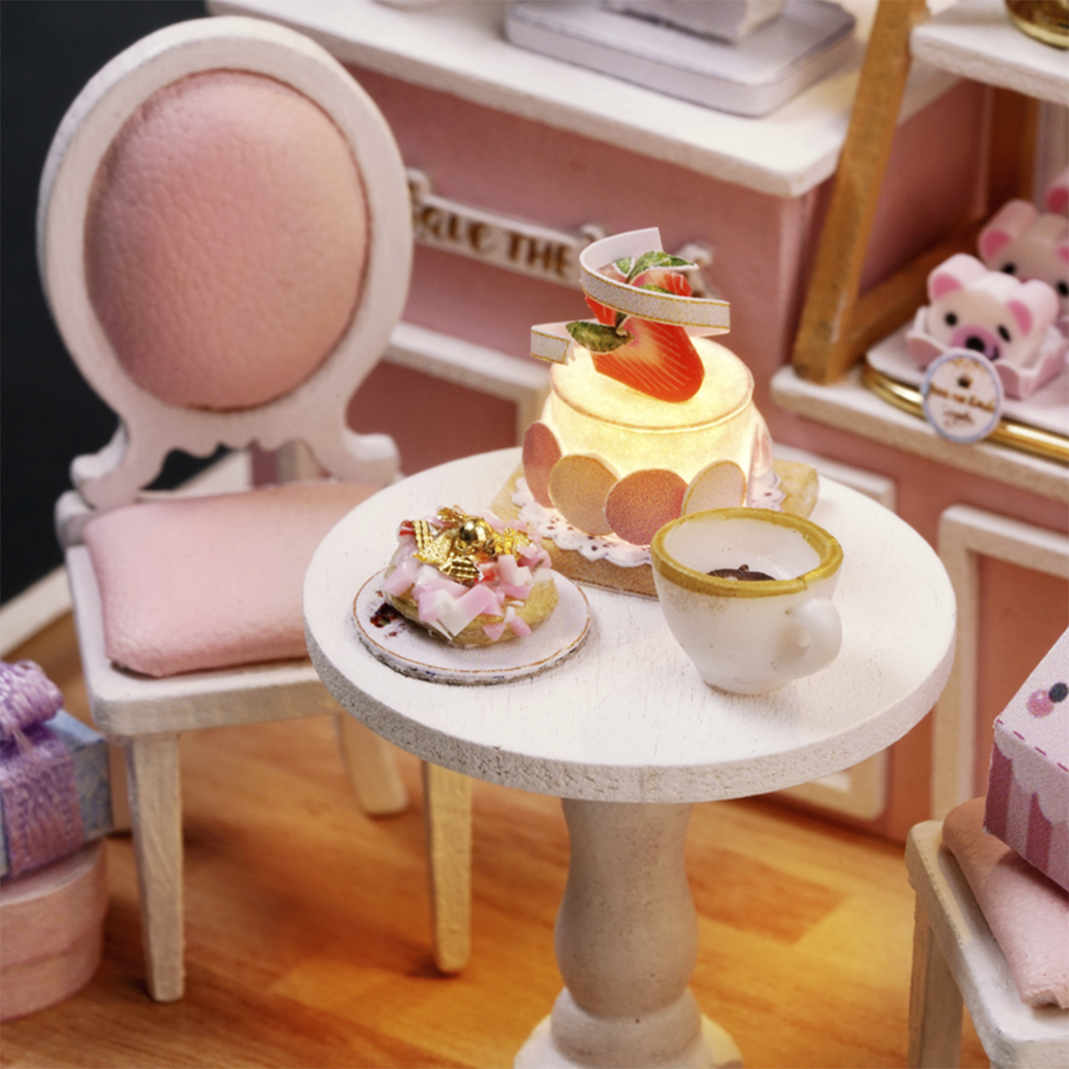Scena in miniatura con tavolo, sedia, torta e tazza da tè. Schema di colori rosa e bianco, con orsacchiotto e confezione regalo.