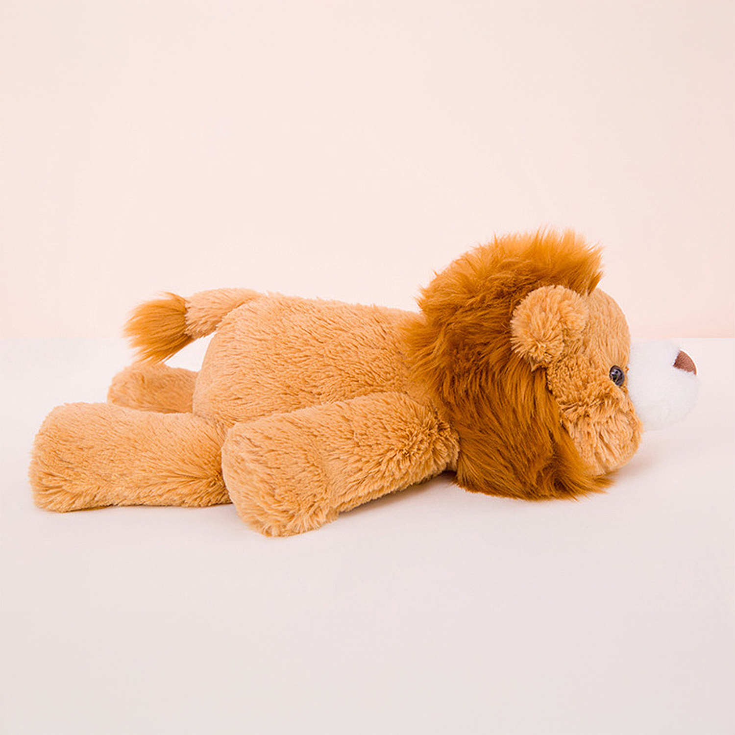 Un leone di peluche sdraiato su una superficie bianca. Ha la pelliccia marrone e una criniera. Lo sfondo è rosa chiaro.