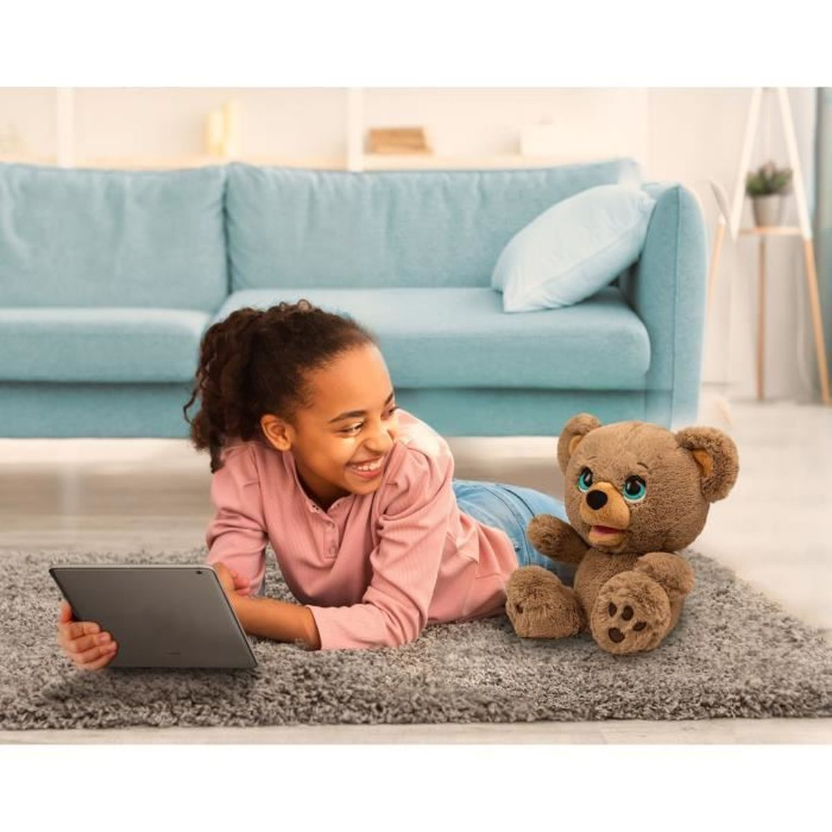 Ein Mädchen lächelt, während es ein Tablet benutzt, auf einem Teppich liegend mit einem Teddybären. Im Hintergrund ist ein blaues Sofa.