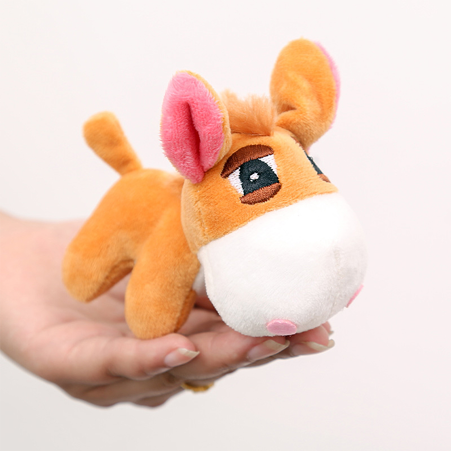 Un piccolo peluche di asino arancione con orecchie foderate di rosa è tenuto in mano a una persona su sfondo bianco.