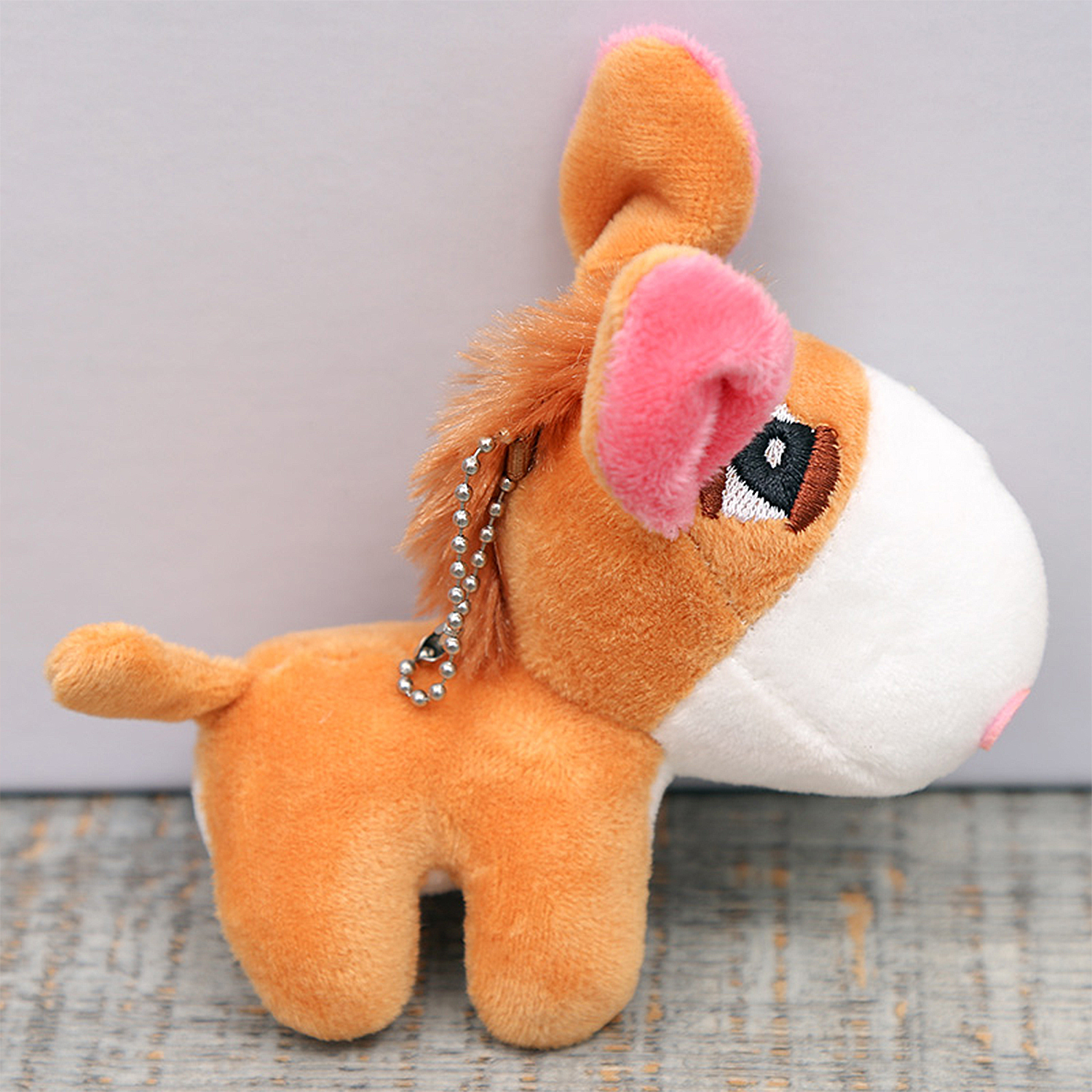 Un peluche a forma di asino. L'asino è arancione con la faccia bianca, le orecchie rosa e una catena d'argento. È su uno sfondo sfocato.