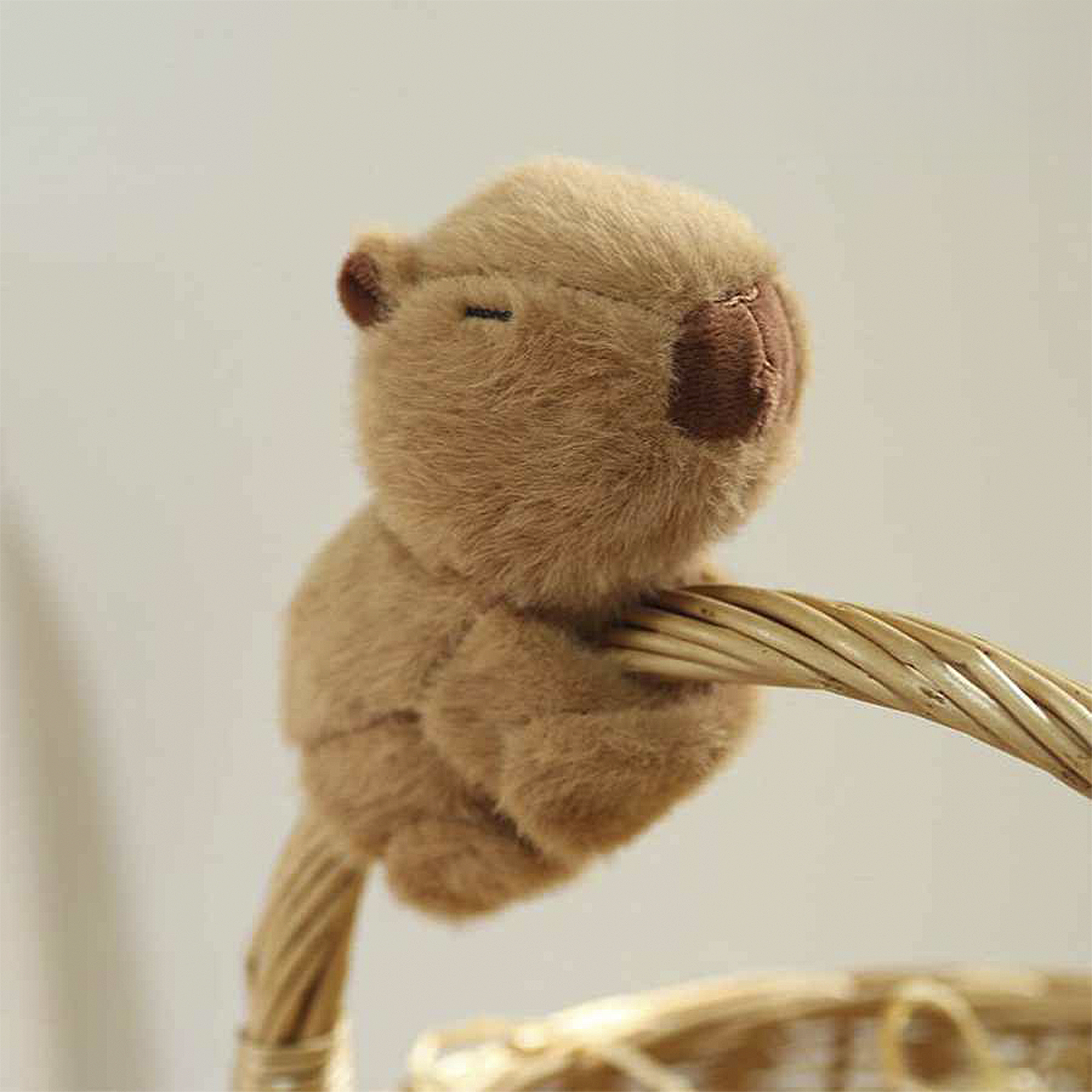 Un capibara de peluche se sienta en una cesta tejida. Es marrón y esponjoso, con un hocico marrón más oscuro.