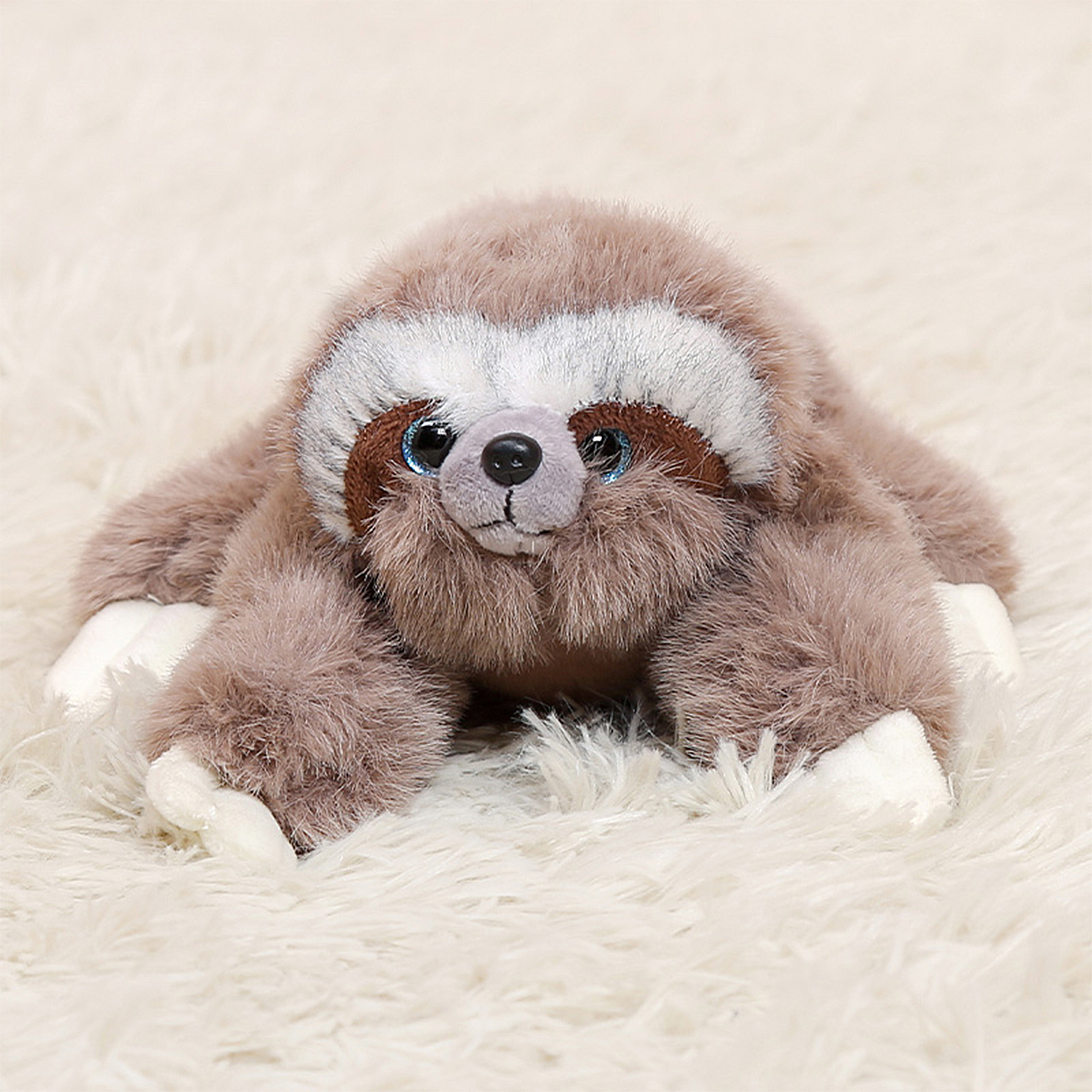 Un giocattolo di peluche a forma di bradipo con pelliccia marrone, artigli bianchi e occhi blu, sdraiato su una superficie bianca e soffice.