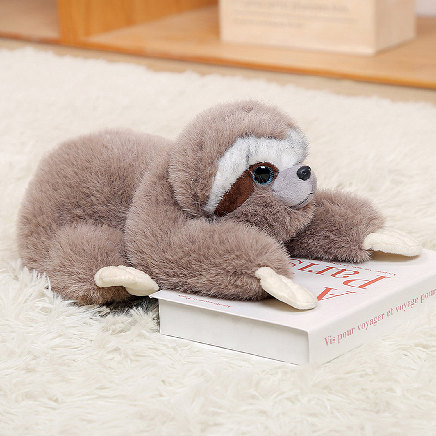 Un peluche di bradipo giace su un soffice tappeto bianco, appoggiando le braccia su un libro. Il bradipo è marrone e beige, con un viso bianco e grandi occhi.