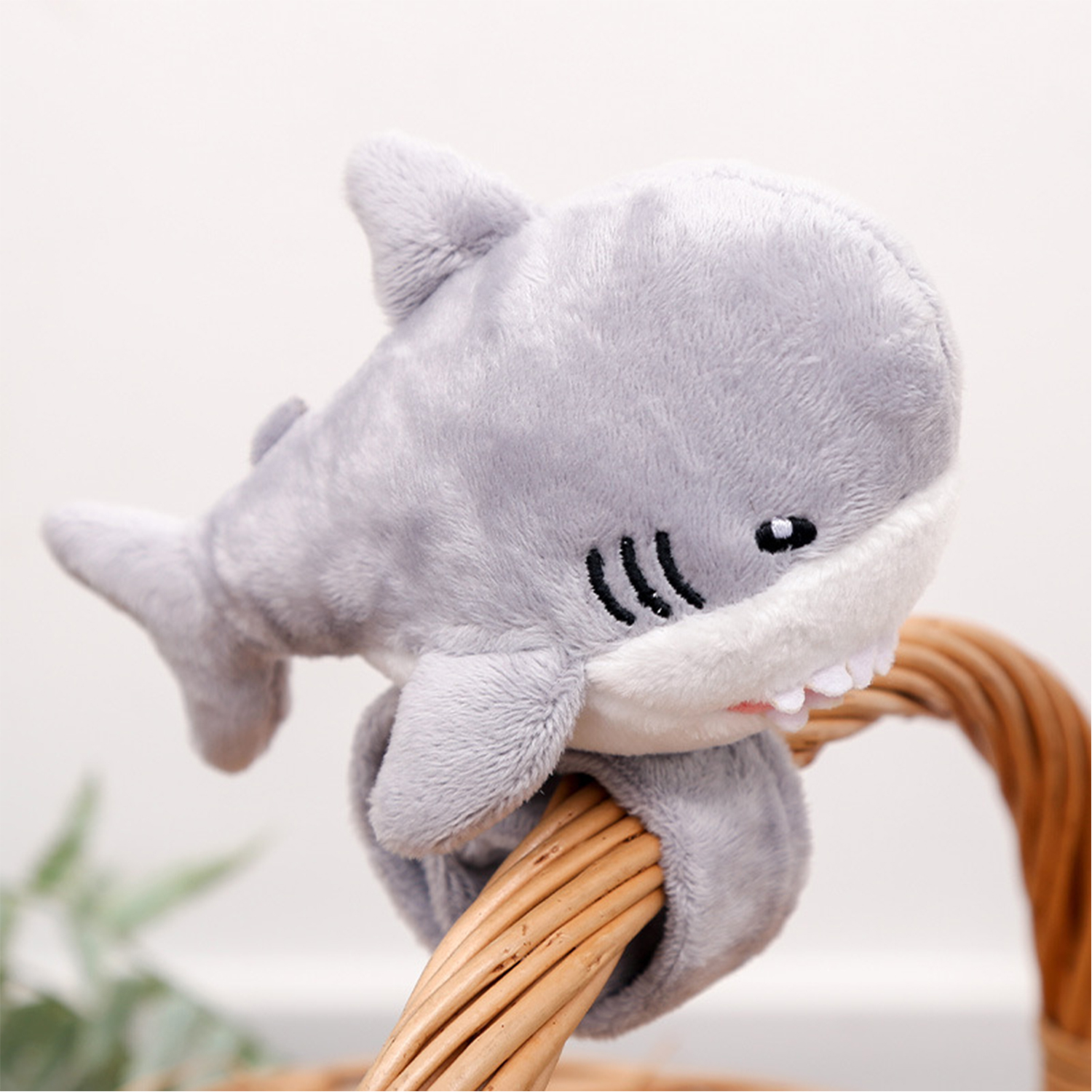 Un giocattolo a forma di squalo di peluche grigio è appollaiato su un cestino di vimini. Lo squalo ha denti bianchi e pancia bianca. Lo sfondo è sfocato.