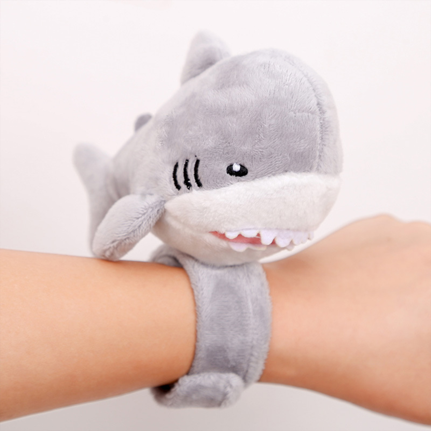 Un braccialetto a forma di squalo di peluche grigio è sul polso di una persona. Lo squalo ha denti bianchi e una pancia bianca. Lo sfondo è bianco.
