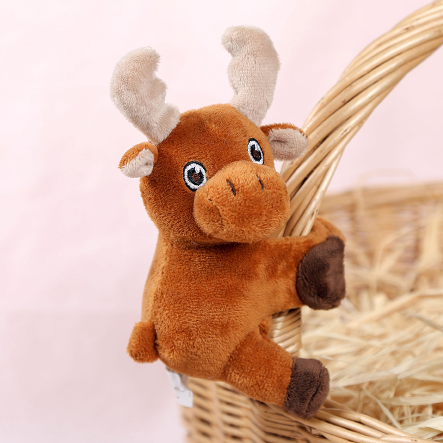 Un alce di peluche marrone si aggrappa a un cestino di vimini. Lo sfondo è rosa e il cestino contiene fieno.