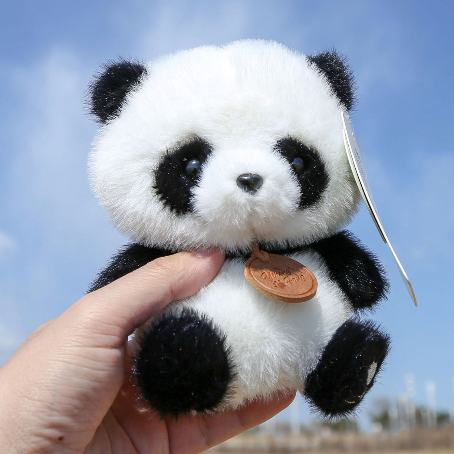 Una mano tiene un panda di peluche con pelliccia bianca e nera, un'etichetta marrone, contro un cielo blu.
