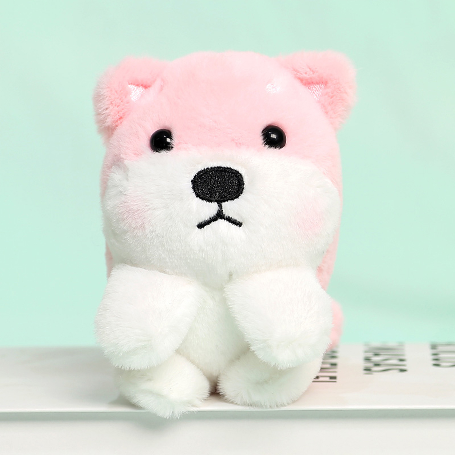 Un giocattolo di peluche a forma di cane rosa e bianco con occhi e naso neri, seduto su una superficie bianca su sfondo verde chiaro.