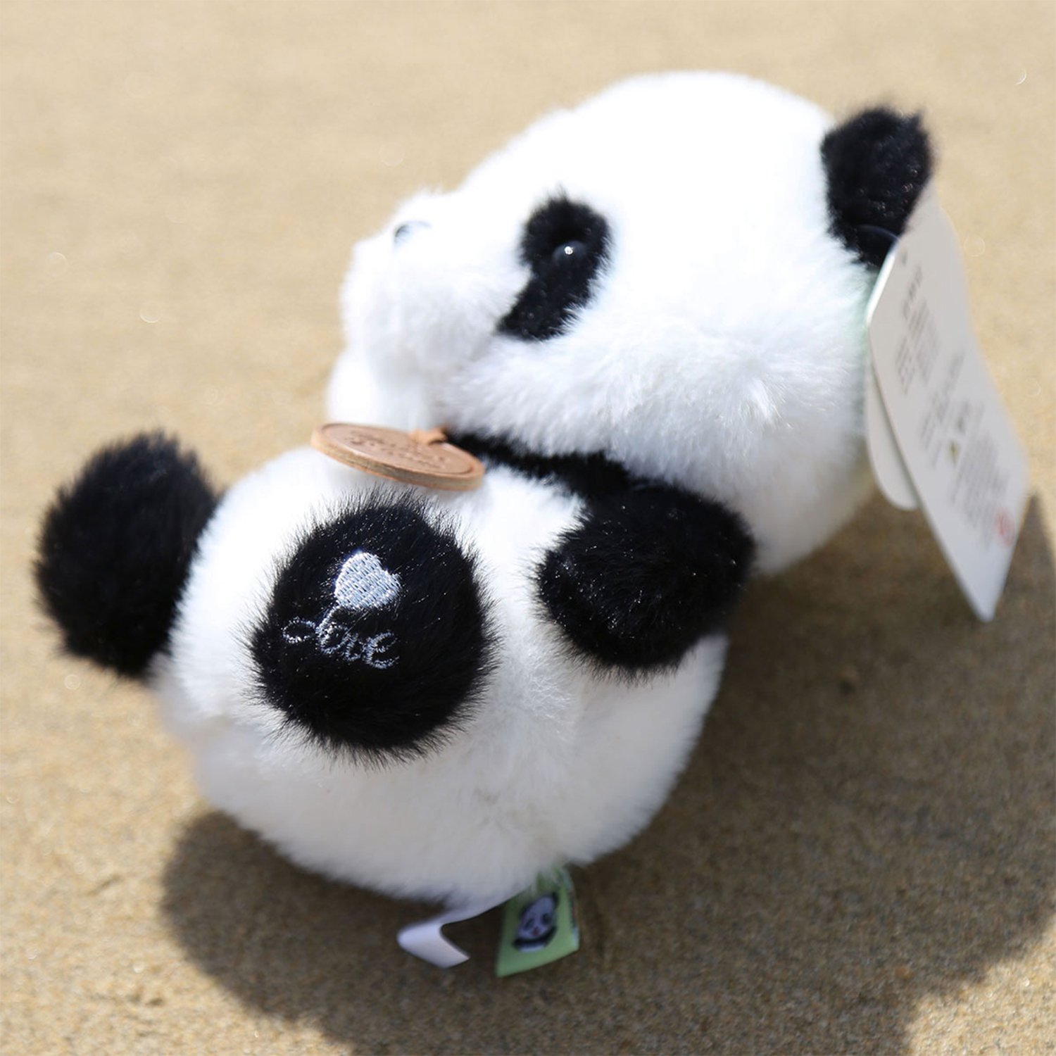 Due peluche a forma di panda con pelo bianco e nero, uno con un cuore e la parola amore sull'occhio. Sono sulla sabbia.