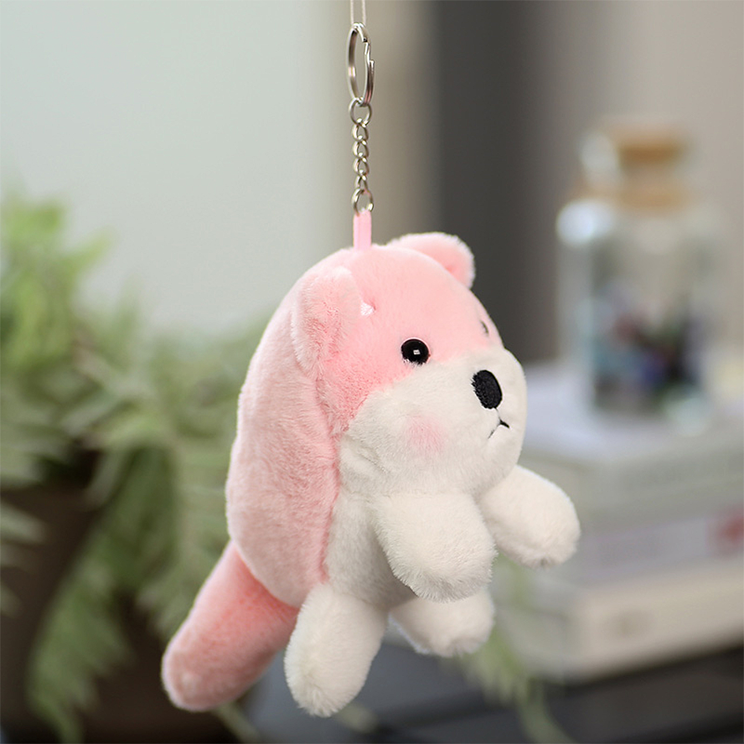 Un portachiavi a forma di animale di peluche rosa e bianco. Ha naso e occhi neri, guance rosee ed è appeso a una catena di metallo.