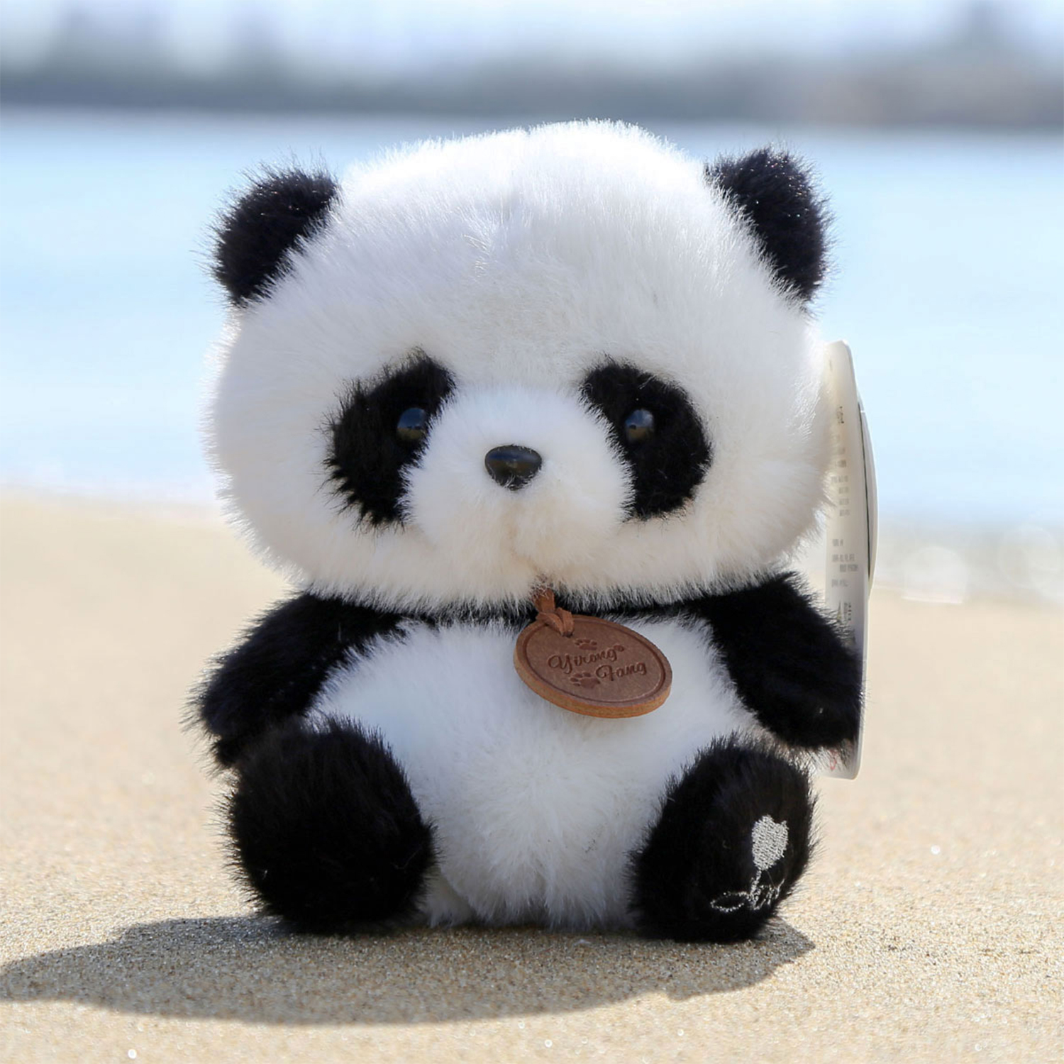Un peluche a forma di panda sulla spiaggia. Ha pelliccia bianca e nera, un'etichetta marrone ed è seduto sulla sabbia con il mare sullo sfondo.