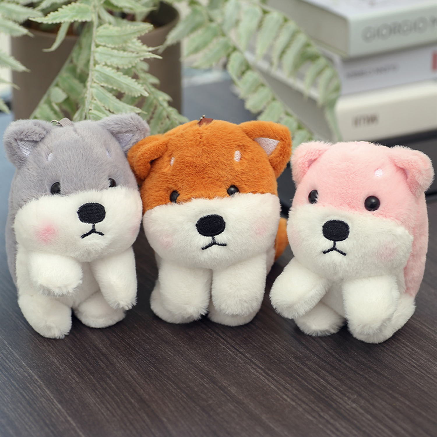 Tre giocattoli di peluche a forma di cane: grigio, arancione e rosa. Ognuno ha la pancia e la faccia bianche. Sono su una superficie di legno con una pianta dietro.