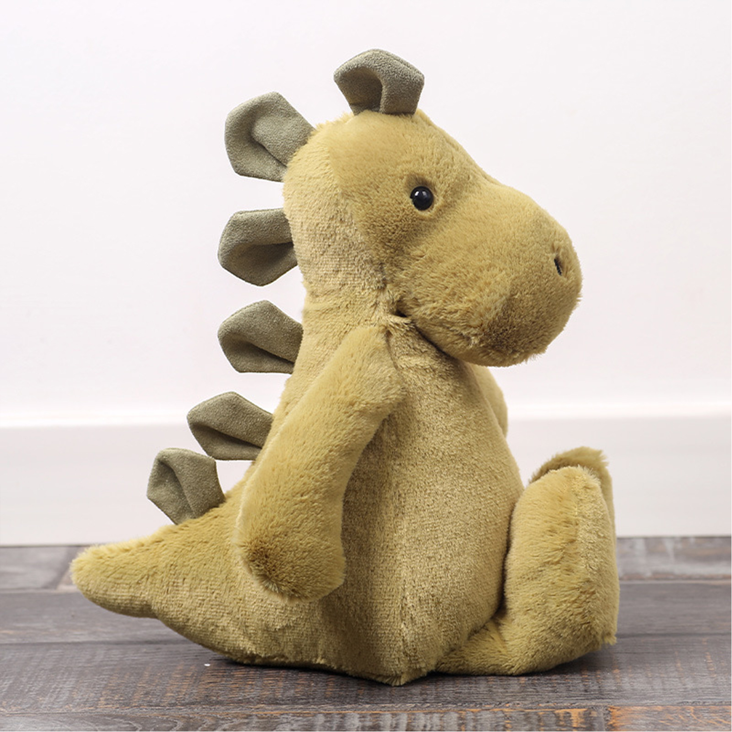 Un peluche di dinosauro morbido, verde-giallo, è seduto su una superficie venata di legno. Ha occhi scuri e spine verde oliva. Lo sfondo è bianco.