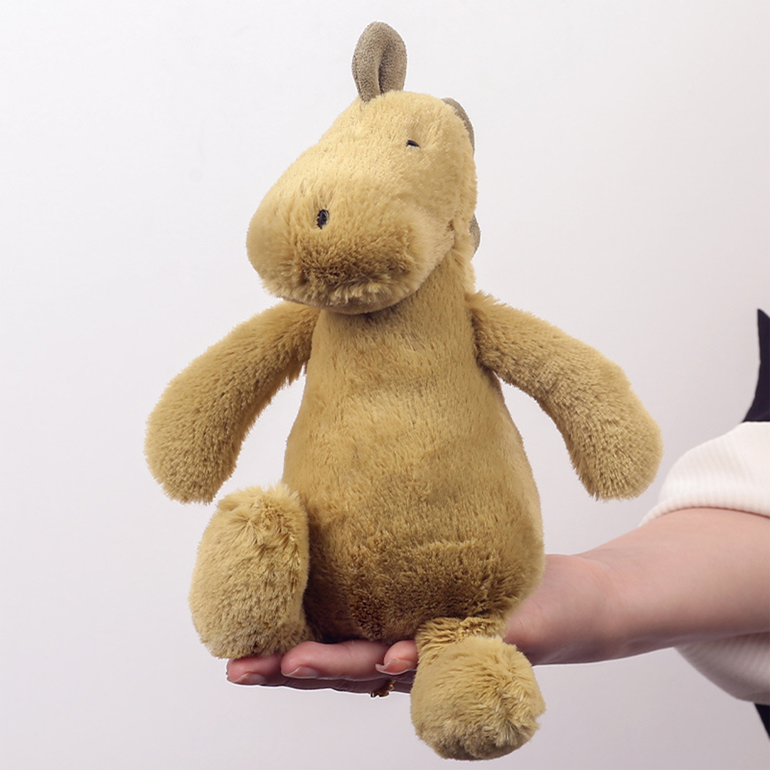 Un peluche marrone chiaro, simile a un cavallo, è tenuto in mano da una persona su sfondo bianco.