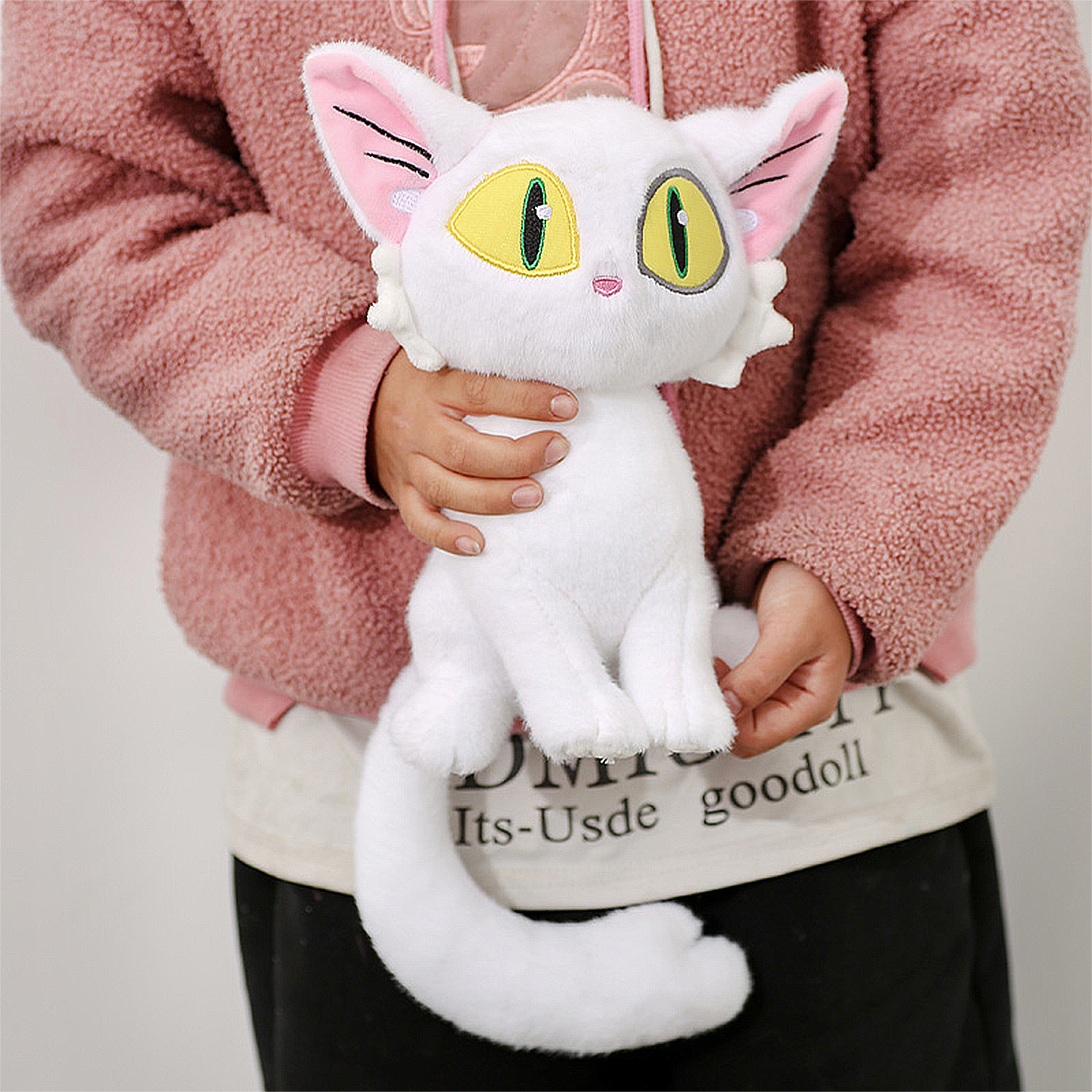 Una persona tiene in mano un peluche di gatto bianco con occhi gialli, orecchie rosa e una lunga coda. La persona indossa una giacca rosa.