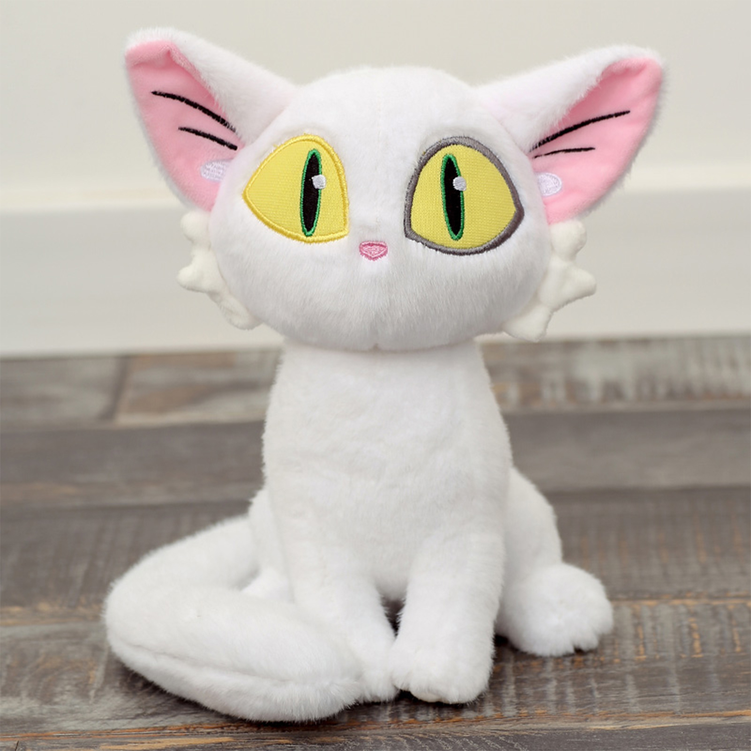 Giocattolo di peluche bianco a forma di gatto con grandi orecchie, occhi gialli e interno orecchie rosa. È su una superficie di legno.
